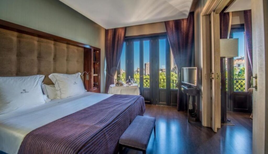 Elegant king room with tall tufted headboard, rich purple drapes, room service table and tree-lined boulevard views