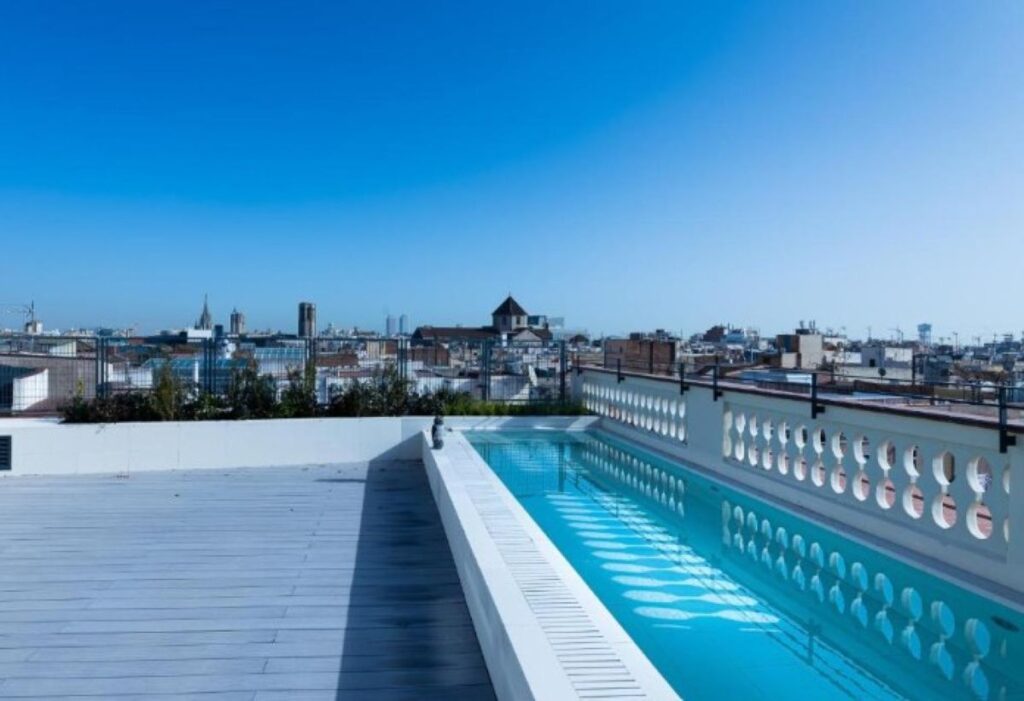 Minimalist rooftop lap pool with ornate white balustrade edging and sweeping 360-degree city rooftop views under blue sky