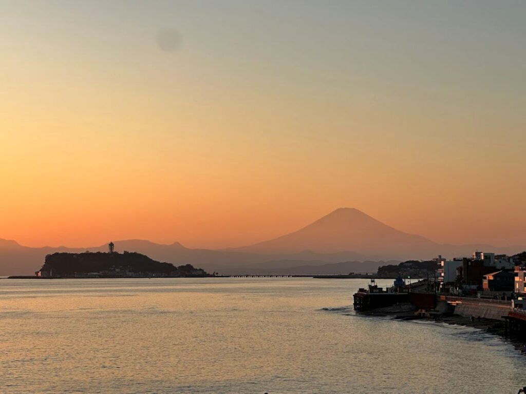 Mount Fuji silhouetted at sunset across the water, with a pier and shoreline buildings in soft orange light.
