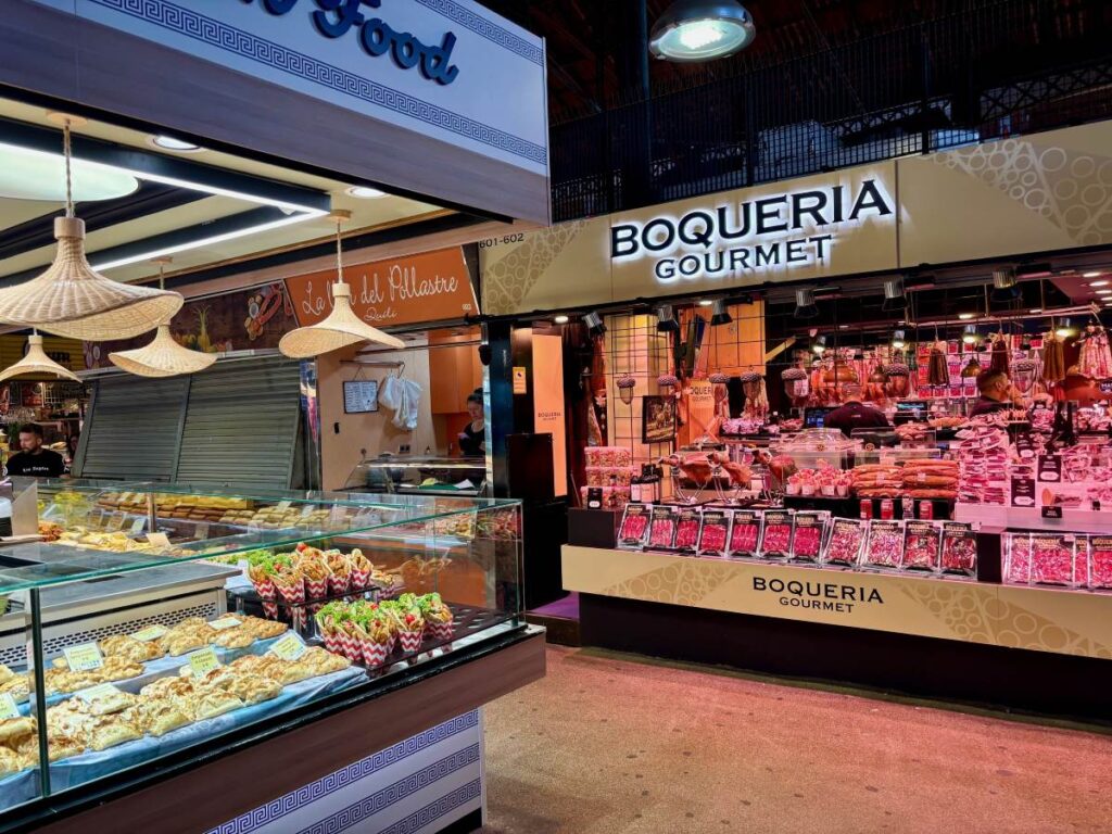 Food stalls inside La Boqueria, with BOQUERIA GOURMET counter, cured meats, and tapas displayed under bright lights.