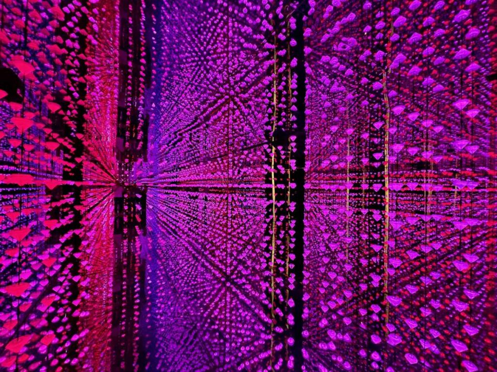 Pink and purple infinity mirror room filled with hanging lights, creating repeating reflections and silhouettes.