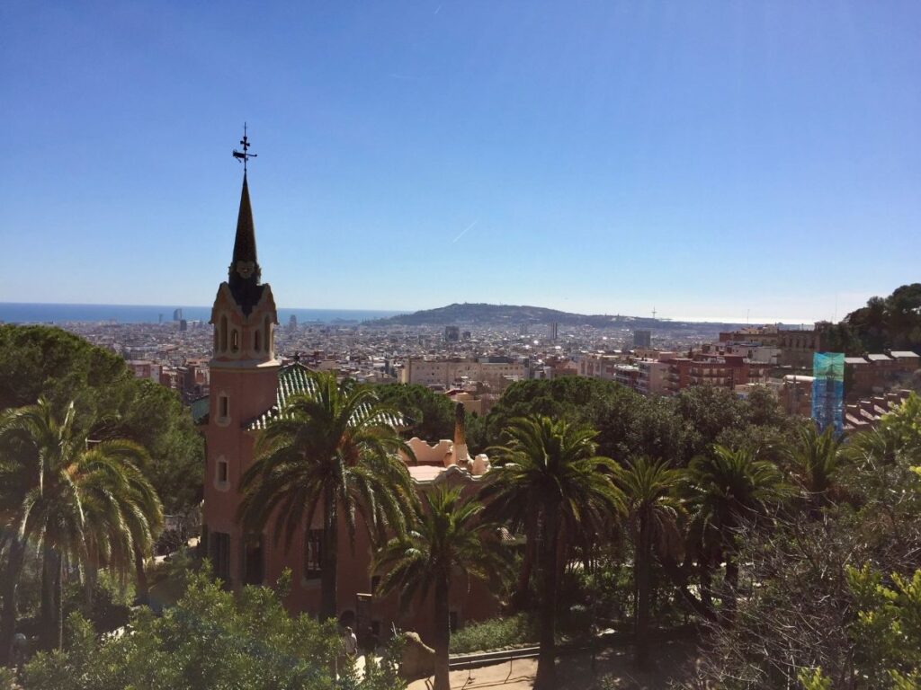 Park Güell viewpoint with palm trees and the fairytale gatehouse tower, overlooking the city and the sea.