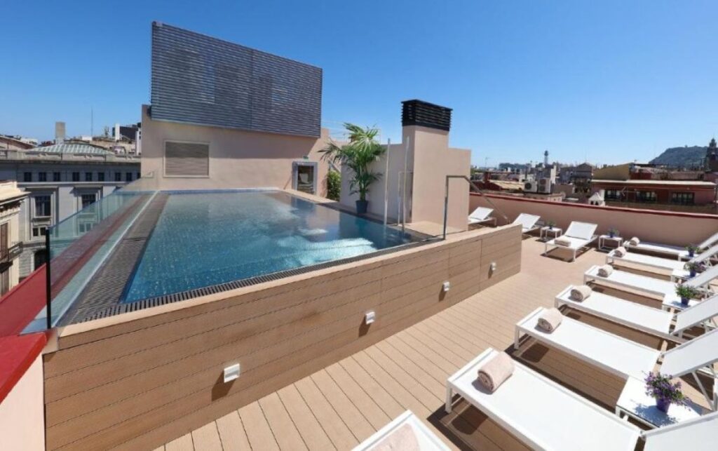 Sunny rooftop infinity pool surrounded by timber decking, white sun loungers and panoramic city rooftop views