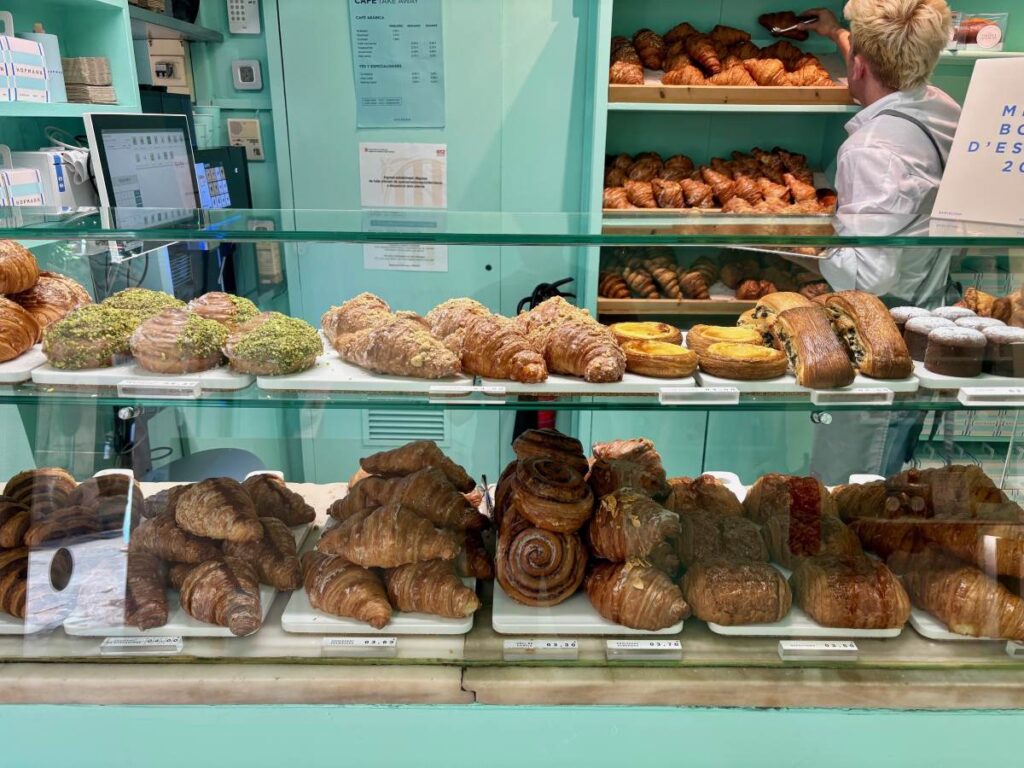 Pastry counter stacked with croissants and buns behind glass, with a baker reaching for fresh trays.