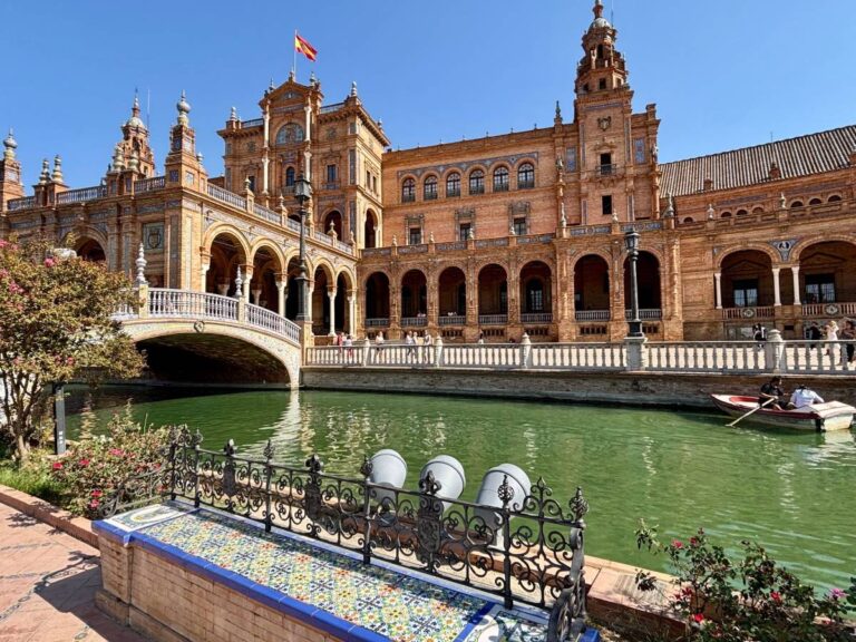 Plaza de España with a tiled bench by the canal and the arched bridge ahead on a sunny day.