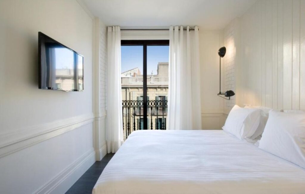 Minimalist white double room with wall-mounted TV, industrial swing arm sconce and ornate iron Juliet balcony