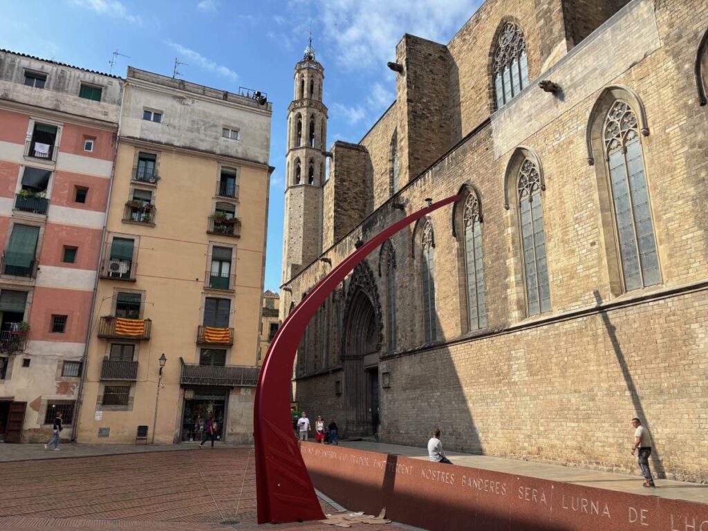 Red curved sculpture beside a Gothic cathedral wall in the Barri Gòtic, with flats and a bell tower in the background.