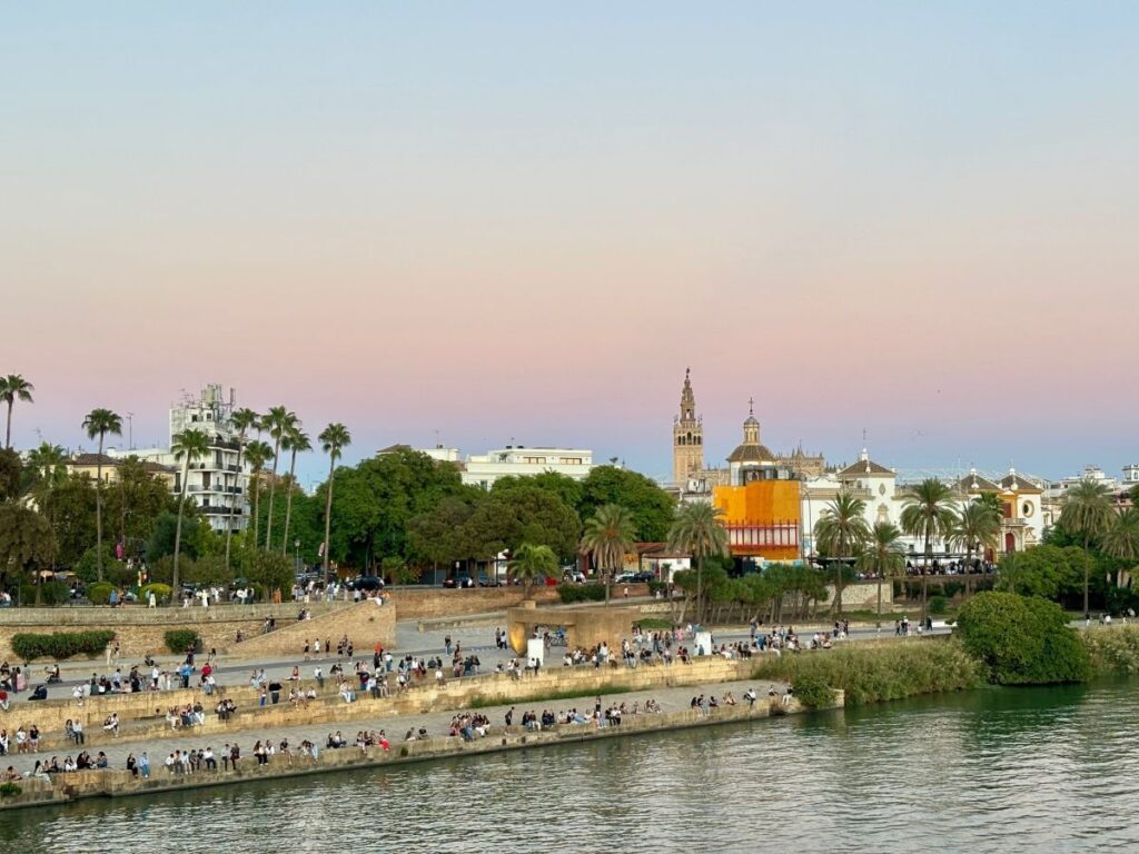 Pastel dusk view over the Guadalquivir, palm-lined riverbank and the Giralda tower above historic rooftops.