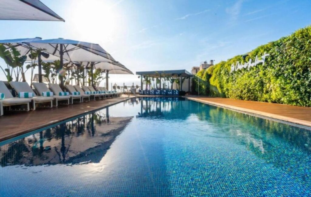 Large outdoor pool with striped sun loungers, white parasols, poolside bar and lush green living wall hedge