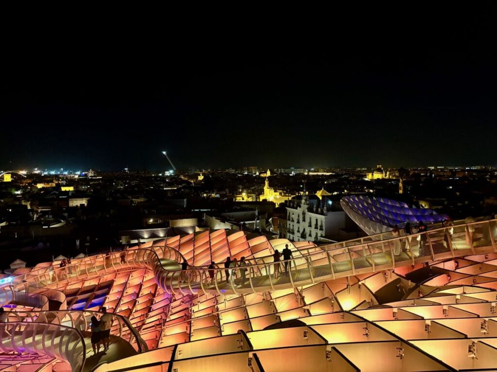 Wide night skyline from Metropol Parasol, glowing walkways and city lights stretching into the distance.