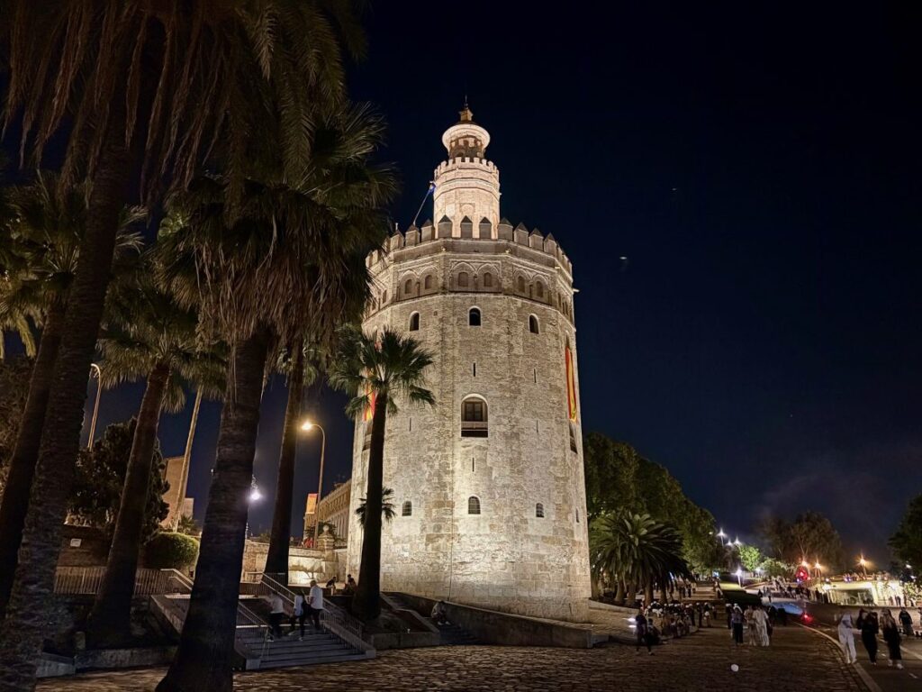 Illuminated Torre del Oro at night, framed by palm trees, with people strolling along the riverside promenade.