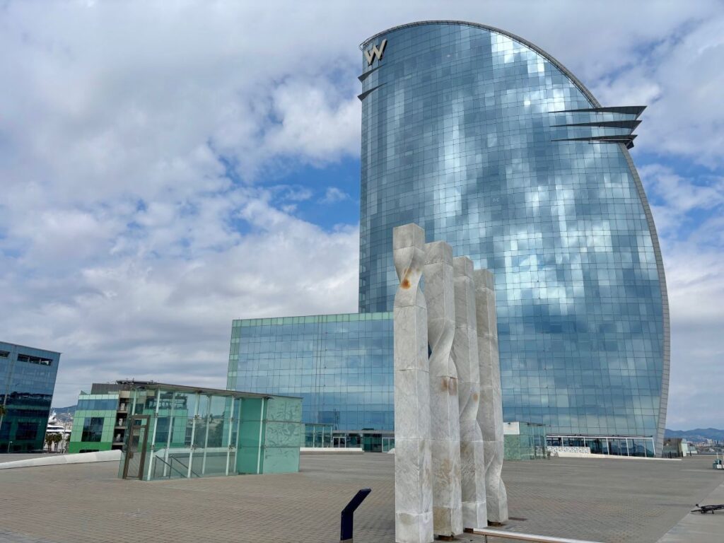 W Barcelona hotel’s glass sail reflecting clouds, with marble sculptures on the rooftop terrace by the waterfront.