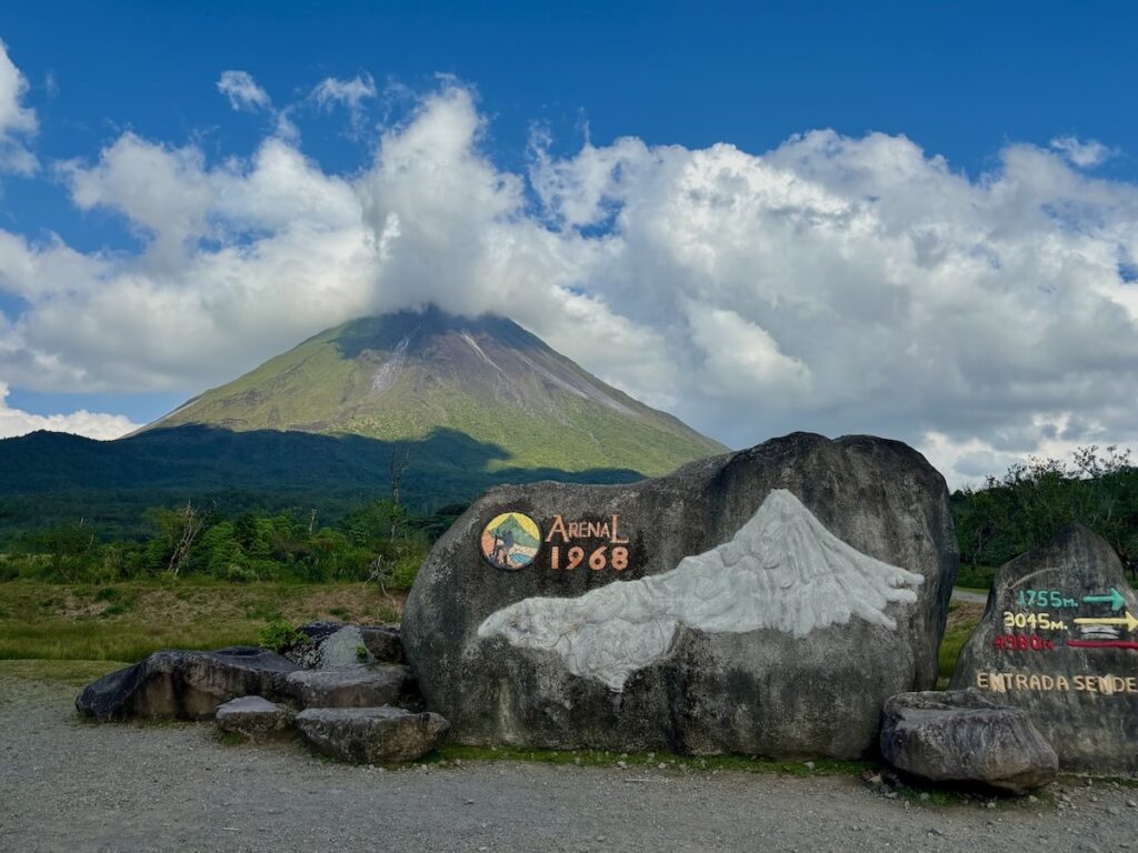 Stone monument with "Arenal 1968" plaque and relief sculpture commemorating the volcanic eruption, with Arenal Volcano and clouds in the background.