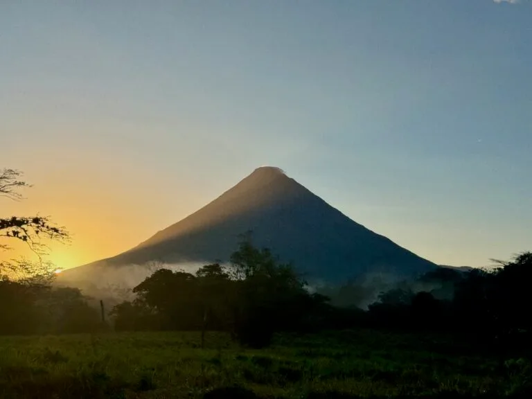 Silhouette of the symmetrical cone of Arenal Volcano against a soft golden sunrise sky, with misty jungle and grassy field in the foreground.