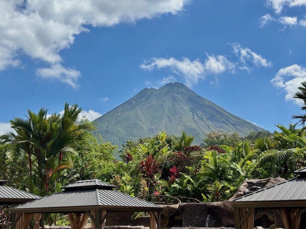 Arenal Volcano rising above tropical palms and red bromeliads, viewed from a resort's thatched cabanas under a bright blue sky with clouds.