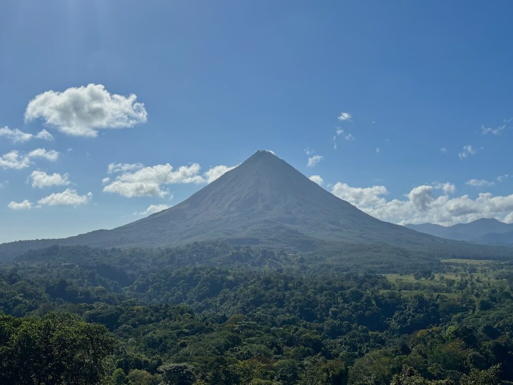 The perfectly conical peak of Arenal Volcano towering over dense green rainforest under a bright blue sky with scattered white clouds. 