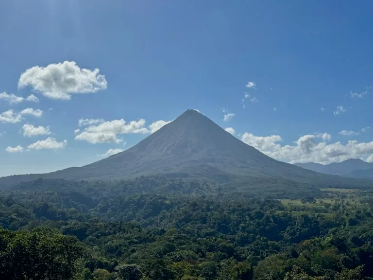 The perfectly conical peak of Arenal Volcano towering over dense green rainforest under a bright blue sky with scattered white clouds.