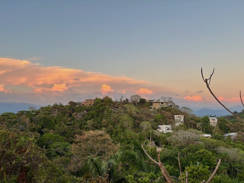 Modern white villas and wooden homes perched on a jungle-covered hillside, framed by a pastel pink and orange sunset over distant mountains.