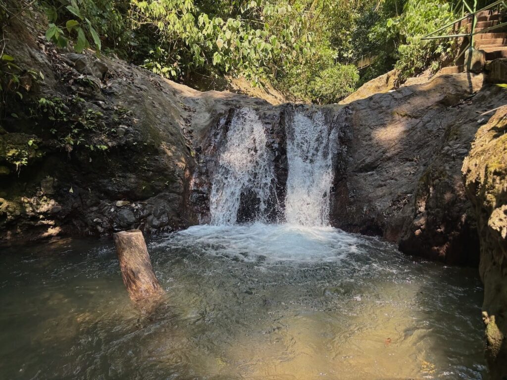 A small twin waterfall tumbling over mossy rocks into a clear natural pool surrounded by lush jungle and a wooden staircase.