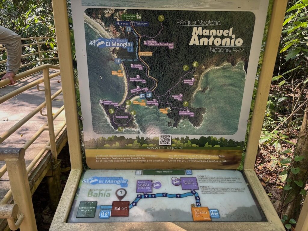 Illustrated signboard map of Manuel Antonio National Park showing trails, beaches and the El Manglar area at the wooden entrance gate.