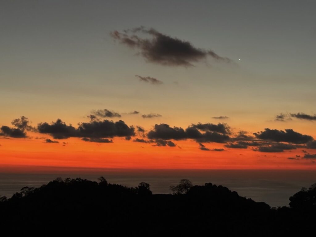 Fiery orange and pink sunset over the Pacific Ocean with a single bright star and silhouetted forested hills in the foreground.