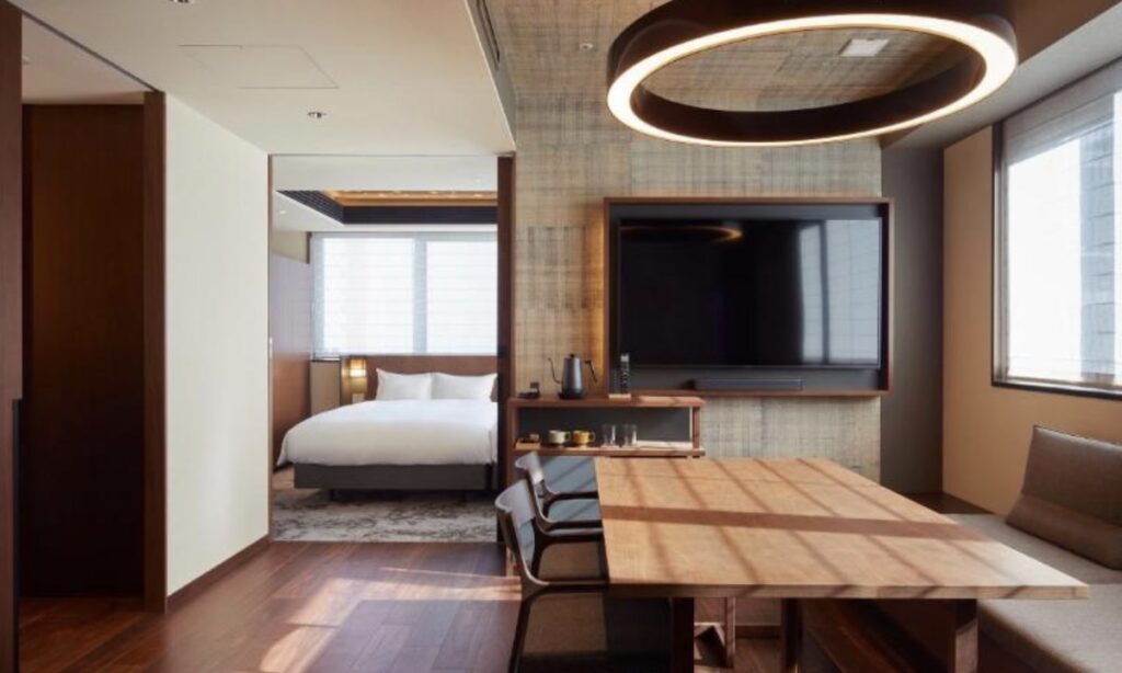 Hotel suite with wooden dining table, circular pendant light and wall-mounted TV, adjoining bedroom with king bed visible through doorwayHotel suite with wooden dining table, circular pendant light and wall-mounted TV, adjoining bedroom with king bed visible through doorway