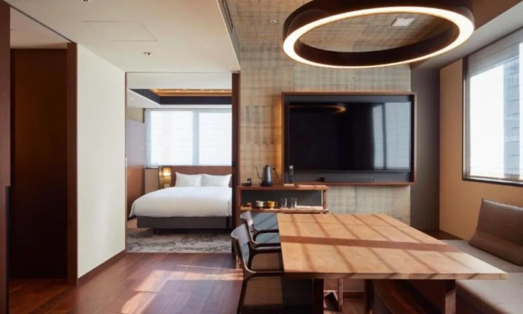 Hotel suite with wooden dining table, circular pendant light and wall-mounted TV, adjoining bedroom with king bed visible through doorwayHotel suite with wooden dining table, circular pendant light and wall-mounted TV, adjoining bedroom with king bed visible through doorway