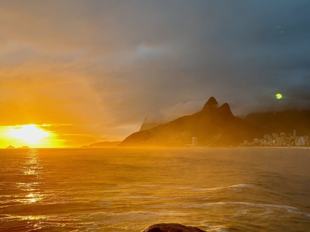 Sunset at Arpoador Rock, golden sun setting behind Two Brothers Mountain, glowing reflection across the Atlantic