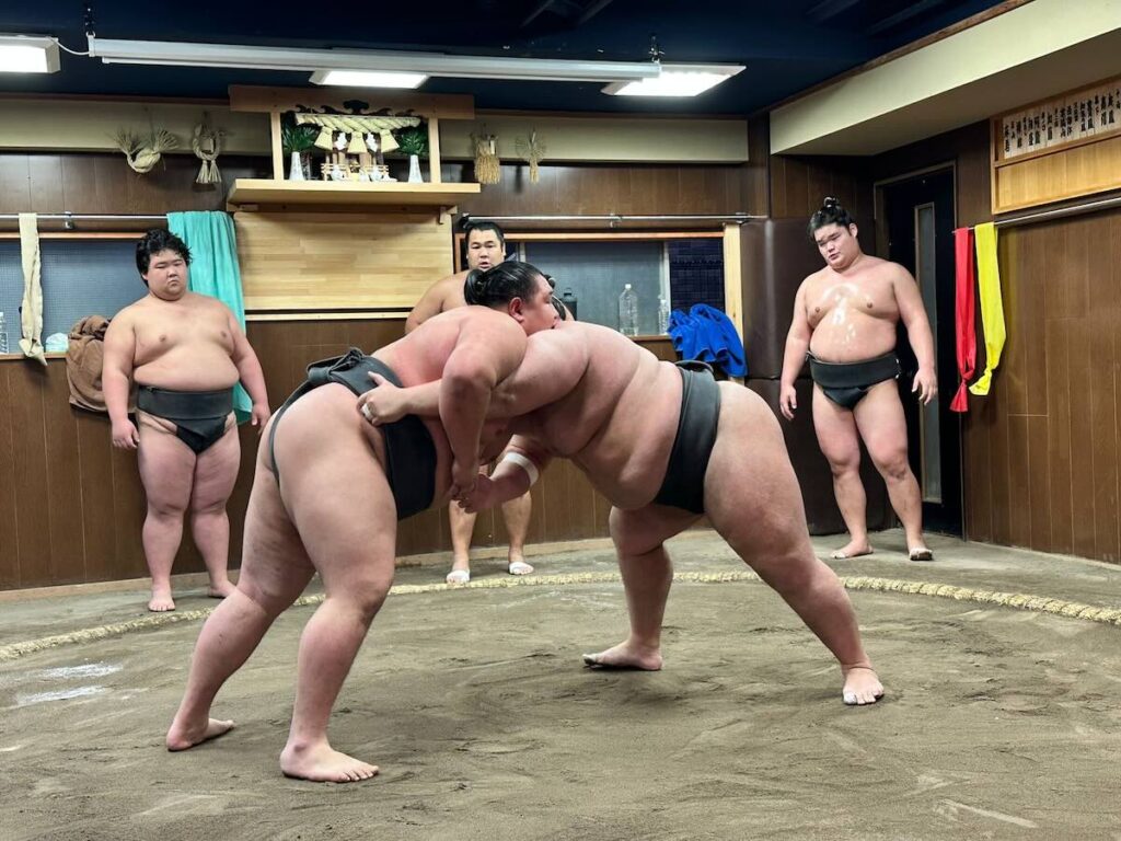 Two sumo wrestlers gripping each other's mawashi belts in a powerful grapple, surrounded by stablemates watching the bout.