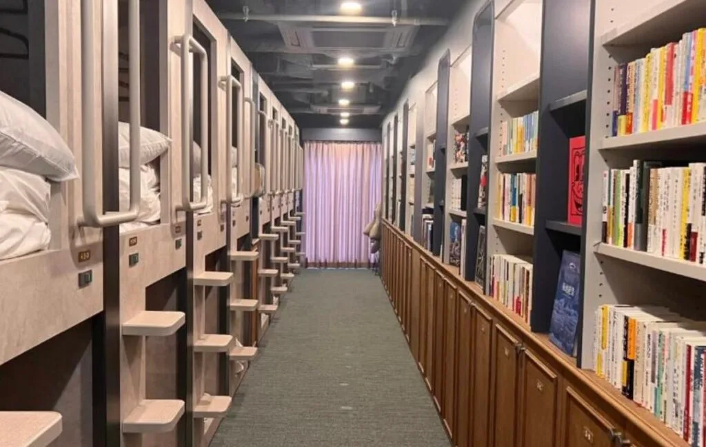 Two-tier capsule pods with ladders facing wall-to-wall bookshelves at BookTeaBed Shibuya capsule hotel