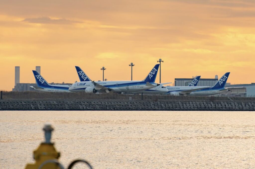 Three ANA aircraft parked on the tarmac at Haneda Airport at sunset, with an orange sky reflecting on the water in the foreground.