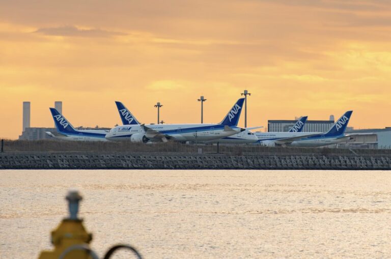 Three ANA aircraft parked on the tarmac at Haneda Airport at sunset, with an orange sky reflecting on the water in the foreground.
