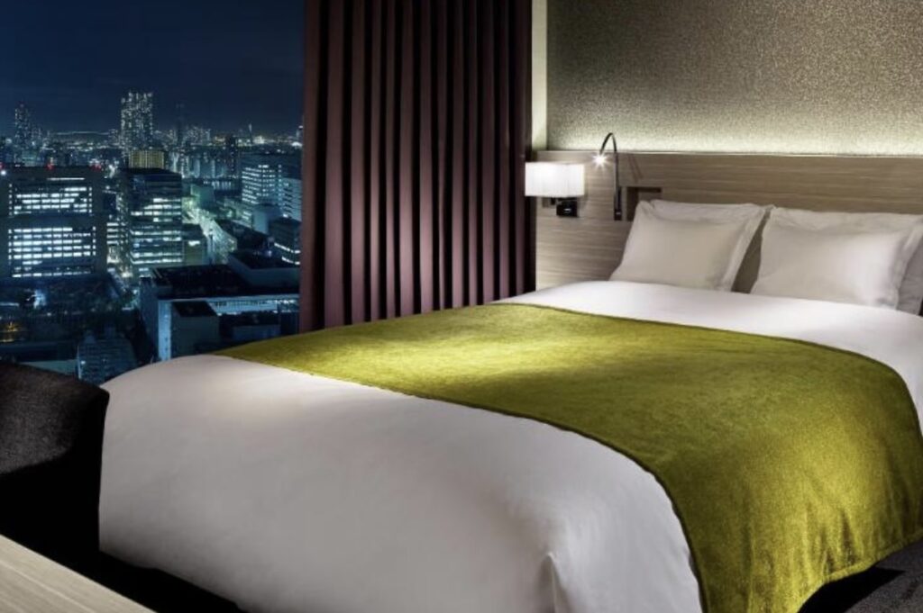 Hotel room with king bed, white linens and olive green throw, beside floor-to-ceiling window showing illuminated city skyline at night