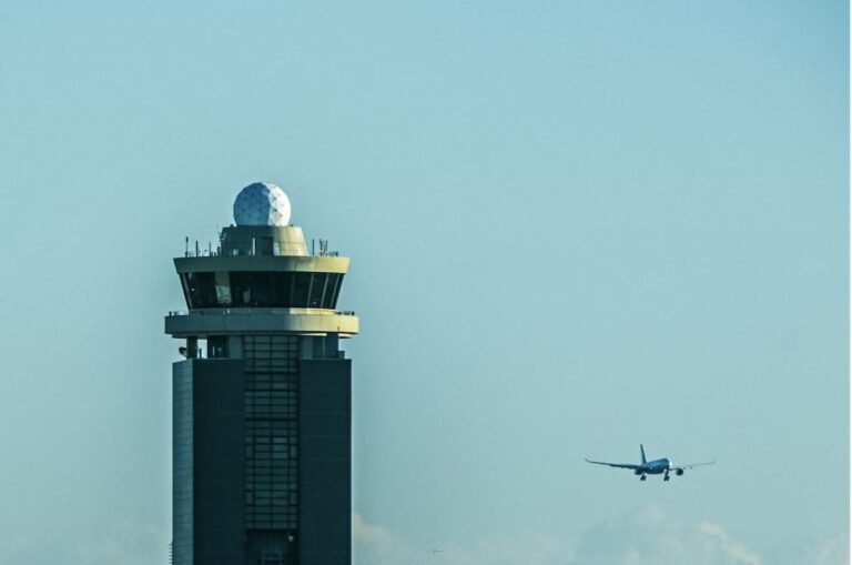 The Narita Airport control tower with its distinctive radar dome against a pale blue sky, as a passenger aircraft approaches to land.