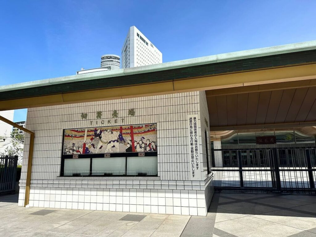 The exterior of the Ryogoku Kokugikan ticket office on a sunny day, with traditional sumo artwork displayed on the tiled façade.