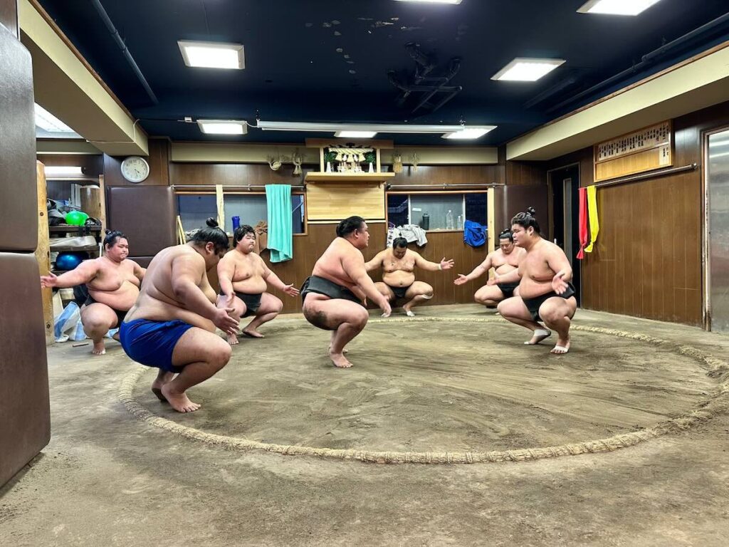Sumo wrestlers in mawashi belts crouching low around the edge of the dohyo training ring during morning practice at a sumo stable.