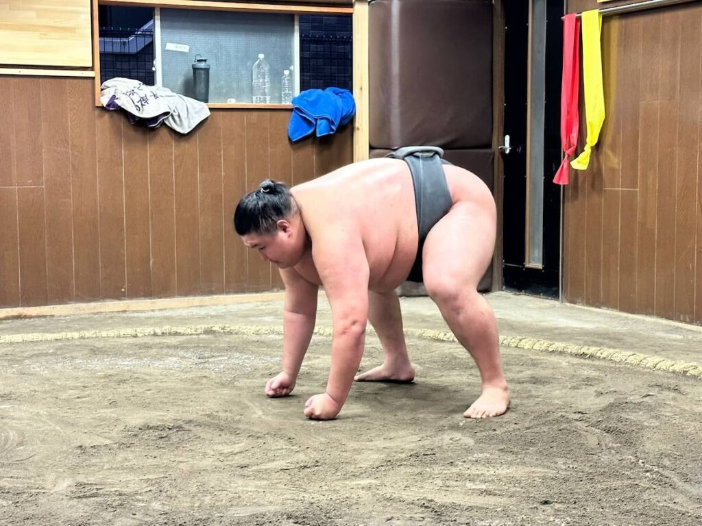 A sumo wrestler in a black mawashi crouching forward with hands on the ring, warming up on the dohyo at a training stable.