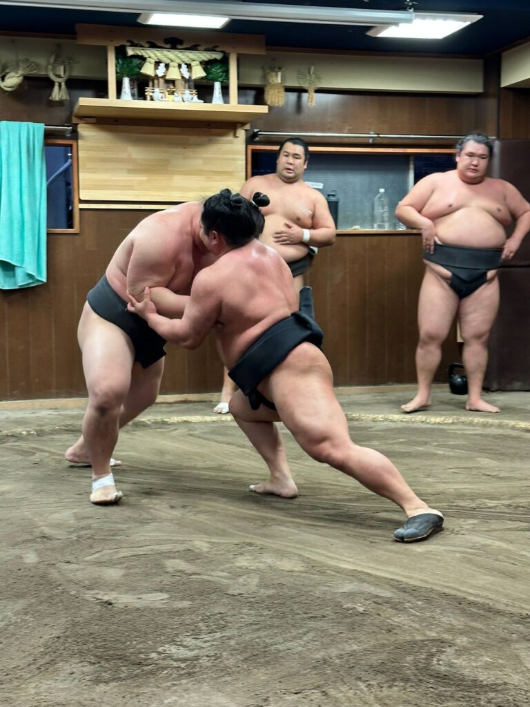 Two sumo wrestlers locked together in a grappling stance on the dohyo, with other wrestlers watching from the side of the training ring.