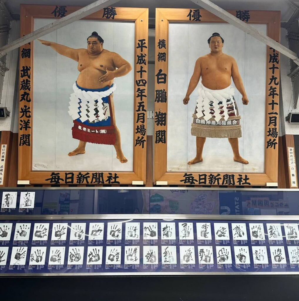 Two large framed yokozuna champion portraits hung above a long display of sumo wrestler handprints and signatures on blue panels.