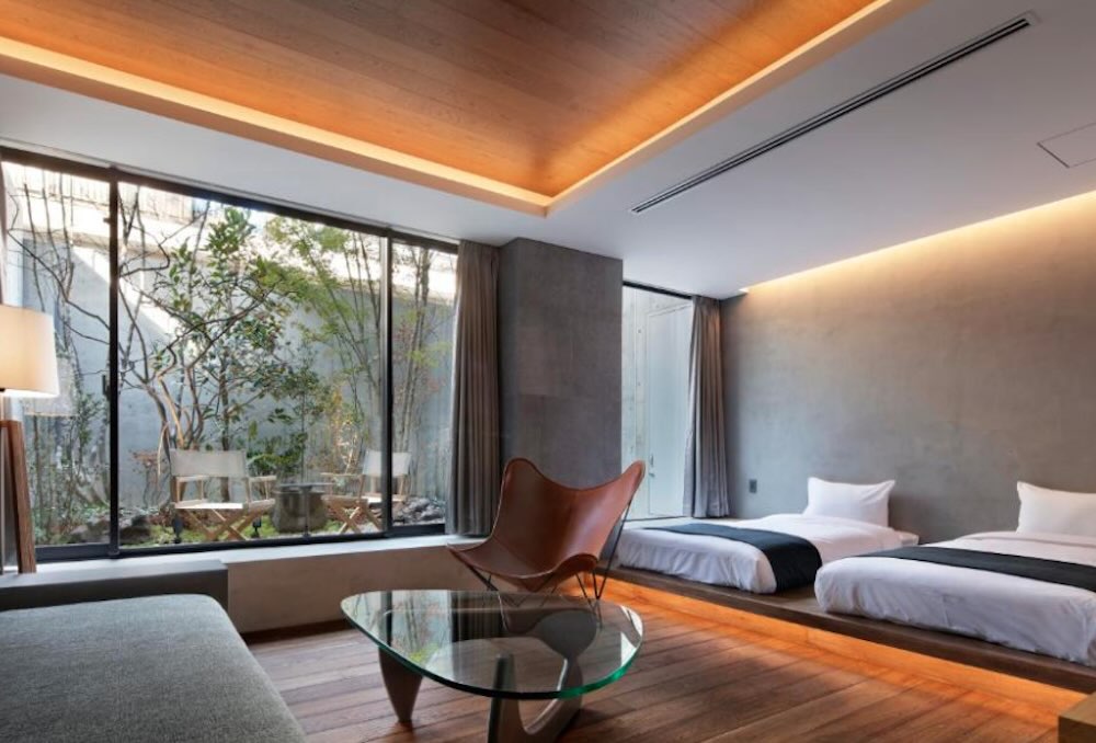 Modern room with two low beds on a raised wood platform, leather chair and big windows onto a small planted courtyard