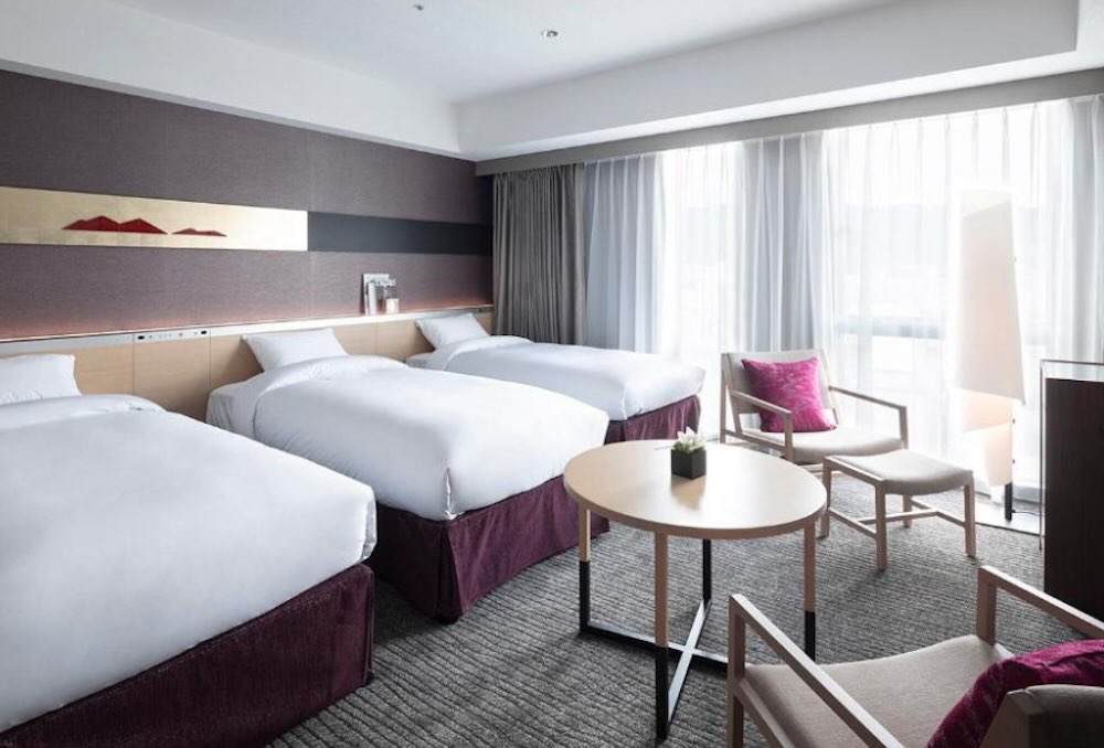 Cross Hotel triple room with three beds, plum-toned base, a round table with armchairs and full-height sheer curtains