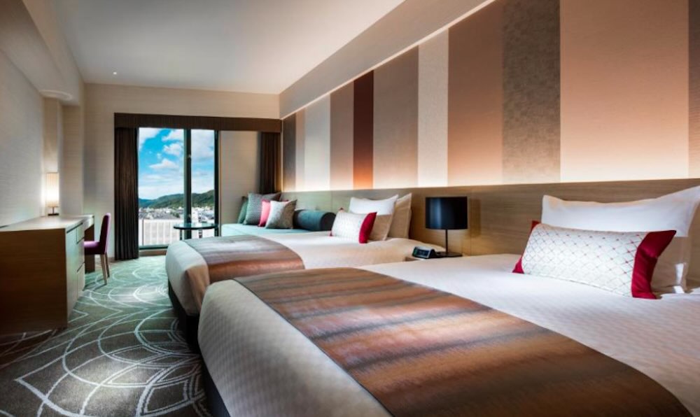 Spacious twin room with two beds, striped feature wall and a balcony window framing hills and the city beyond