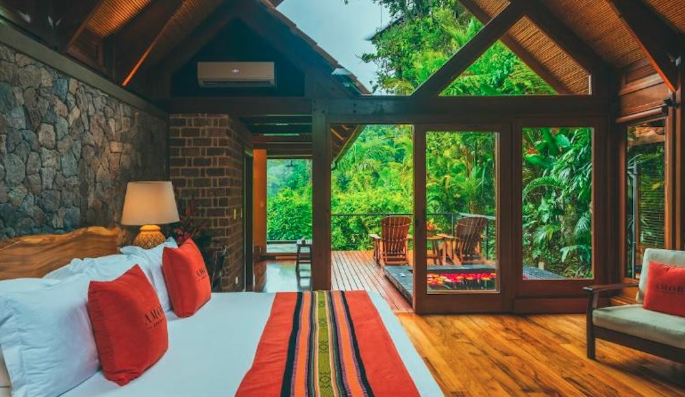 Casita bedroom with a stone wall and pitched timber ceiling, opening through glass doors onto a deck in dense rainforest