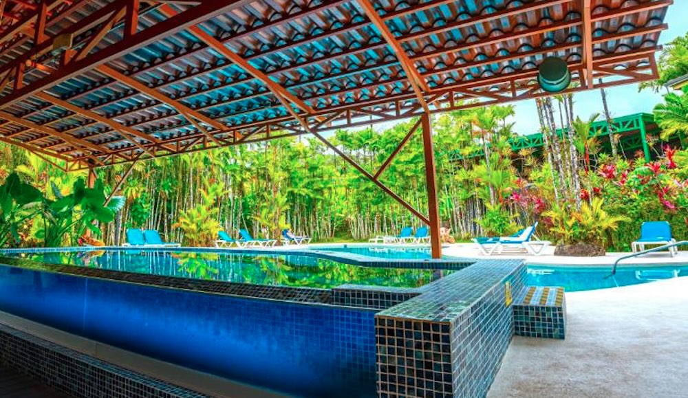 Blue tiled swimming pool under a slatted wooden canopy, with white sun loungers along the edge and dense bamboo planting behind
