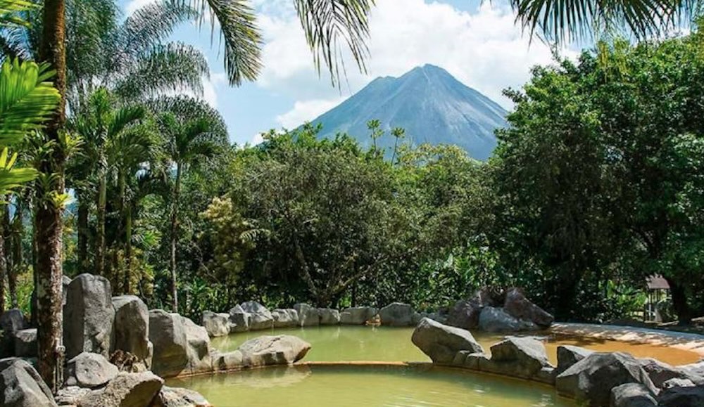 Rock-edged mineral pool with cloudy ochre water, palms and rainforest behind, and Arenal Volcano rising above the treeline