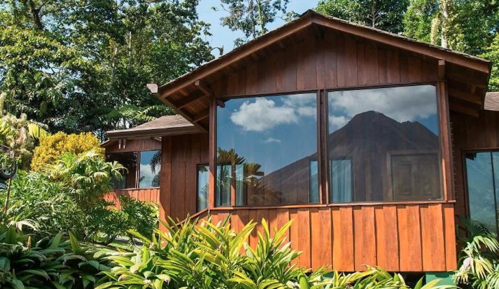 Timber bungalow with a broad picture window reflecting the volcano's cone, framed by ferns and flowering shrubs