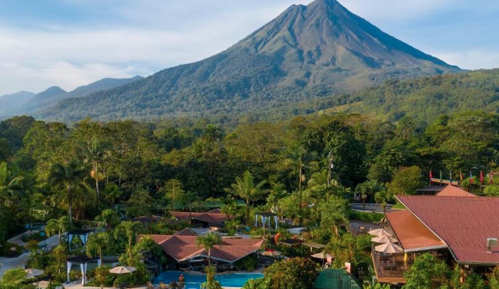 Aerial view of a red-roofed resort with a swimming pool and gardens, backed by rainforest and the cone of Arenal Volcano