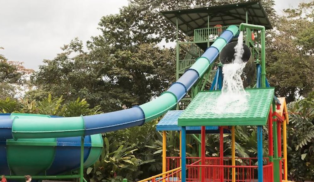 Green and blue tube waterslide curving down from a tower, beside a tipping bucket splash frame in primary colours