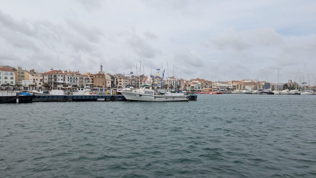 Fishing boats and yachts moored in Cambrils harbour, with the seafront town and church tower under a cloudy sky