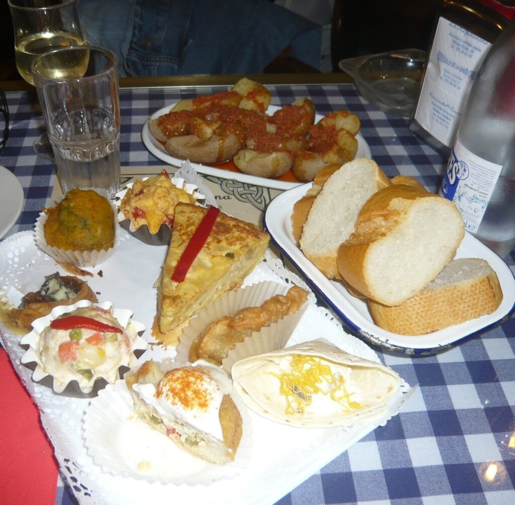 A spread of Spanish tapas including tortilla, croquettes and patatas bravas with bread and white wine on a checked tablecloth
